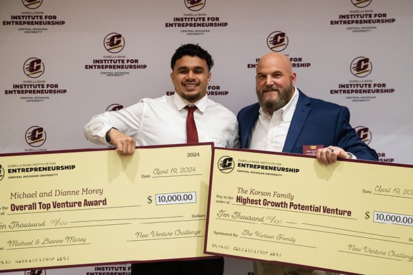 A student standing with a faculty member holding big winning checks from the New Venture Challenge 2024.
