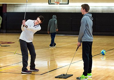 Ben Rollenhagen looks at the golf club he is holding in preparation to swing. Ben is demonstrating to a student who is standing in front of him resting his golf club on a platform on the gym floor.