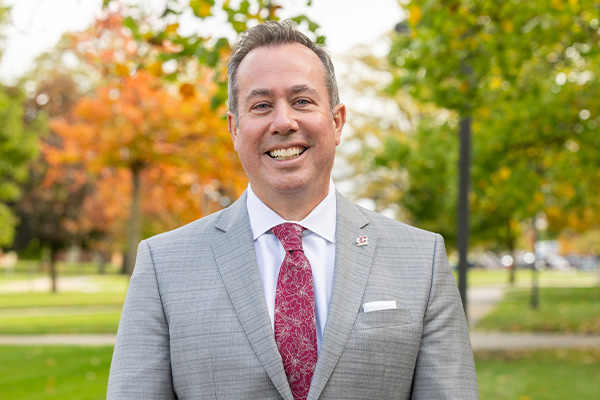 President MacKinnon Headshot 2 Professional headshot of President Neil MacKinnon wearing a gray suit smiling at the camera outdoors with trees in the background.