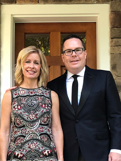 A man with glasses in a black suit, white dress shirt and black tie stands next to a woman in a black and red paisley dress. In the background is a door to a building.